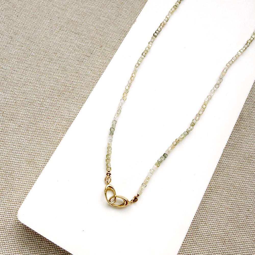 A delicate white zircon gemstone necklace with a gold infinity-style pendant, displayed on a white rectangular card over a beige textured fabric background.