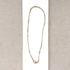 A delicate white zircon gemstone necklace with a gold infinity-style pendant, displayed on a white rectangular card over a beige textured fabric background.