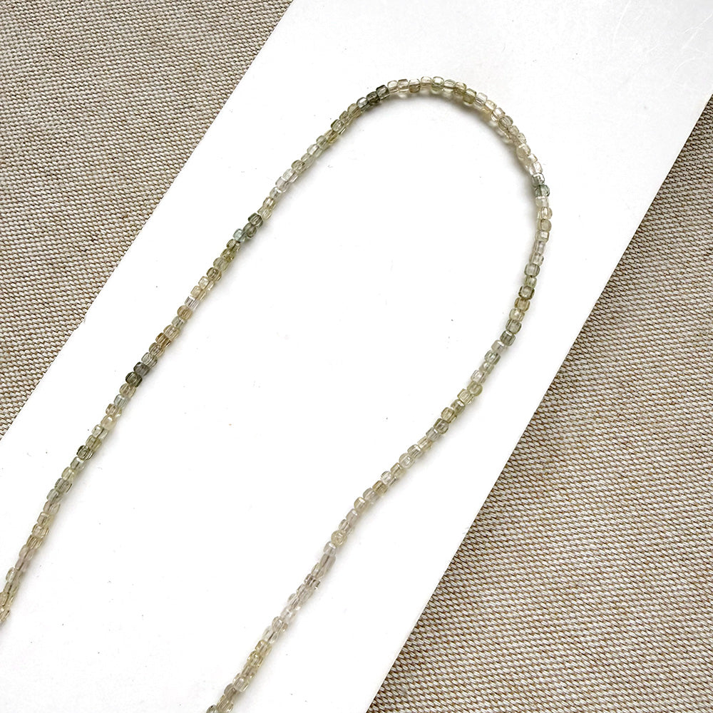 A delicate beaded necklace made of small, faceted gray-green gemstone beads, displayed on a white card over a textured beige fabric background.