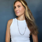 A woman with long blonde-highlighted hair wearing a white ribbed tank top and a long beaded watermelon tourmaline necklace, posing against a dark blue-gray wall.