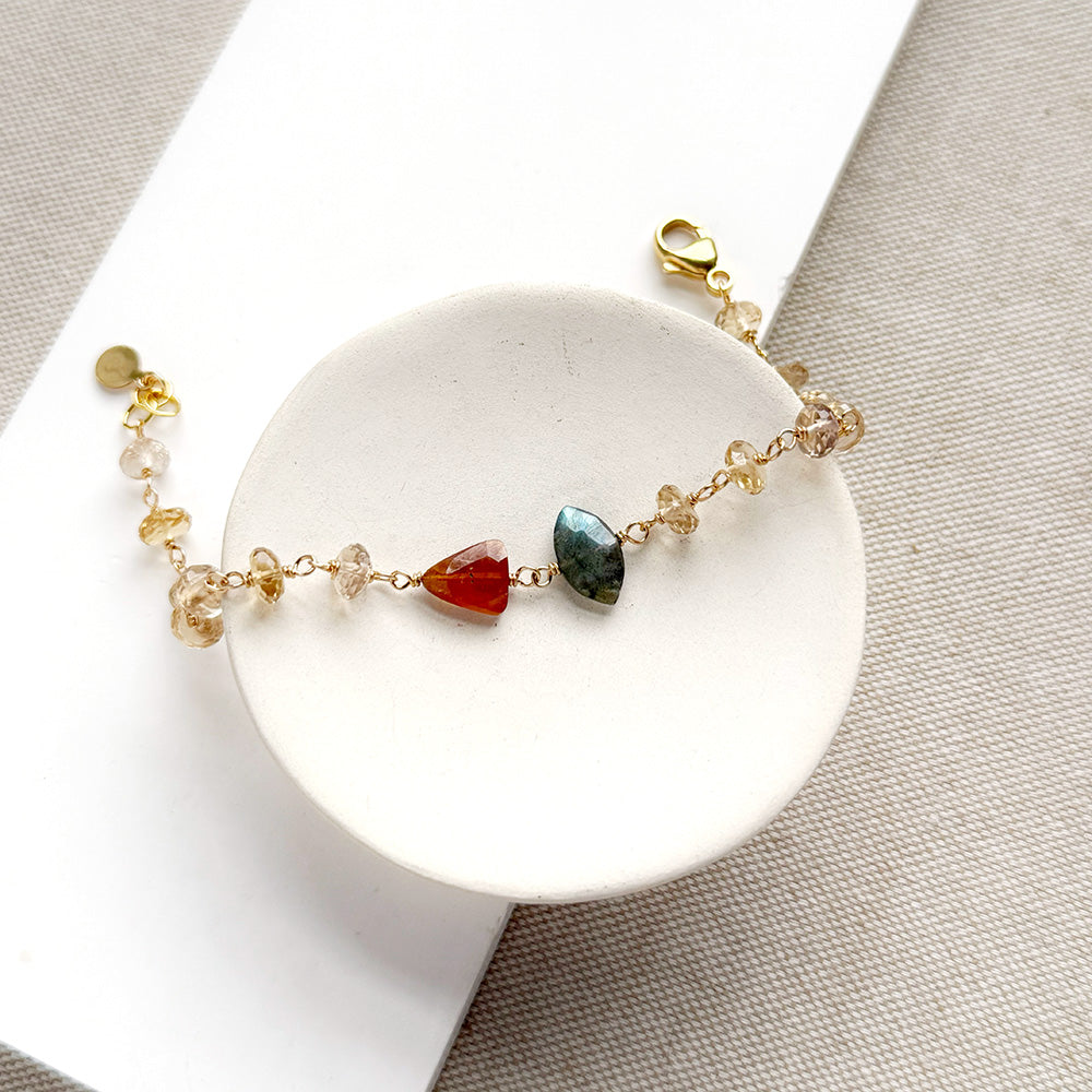 A delicate gold chain bracelet with three central gemstone beads (red, green, and clear) displayed on a white circular stand over a beige textured fabric background.