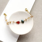 A delicate gold chain bracelet with three central gemstone beads (red, green, and clear) displayed on a white circular stand over a beige textured fabric background.