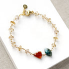 A delicate gold chain bracelet with clear faceted crystal beads and two prominent gemstone charms, one orange-red and one teal-green, displayed on a white card.
