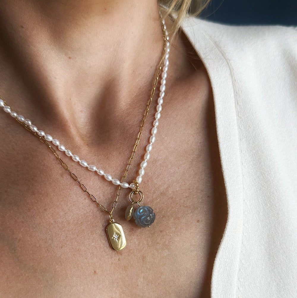 A close-up of a gold chain necklace worn on a person's neck, featuring a small gold tag charm with a star and a round carved labradorite charm.
