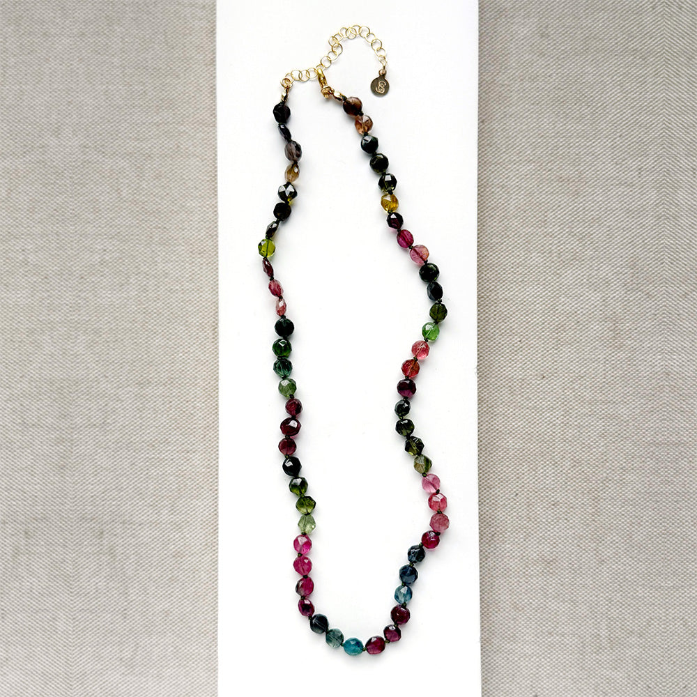 A necklace with faceted Watermelon Tourmaline gemstone beads in a multicolor mix of green, pink, and blue, on a white surface with textured beige fabric background
