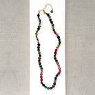 A necklace with faceted Watermelon Tourmaline gemstone beads in a multicolor mix of green, pink, and blue, on a white surface with textured beige fabric background