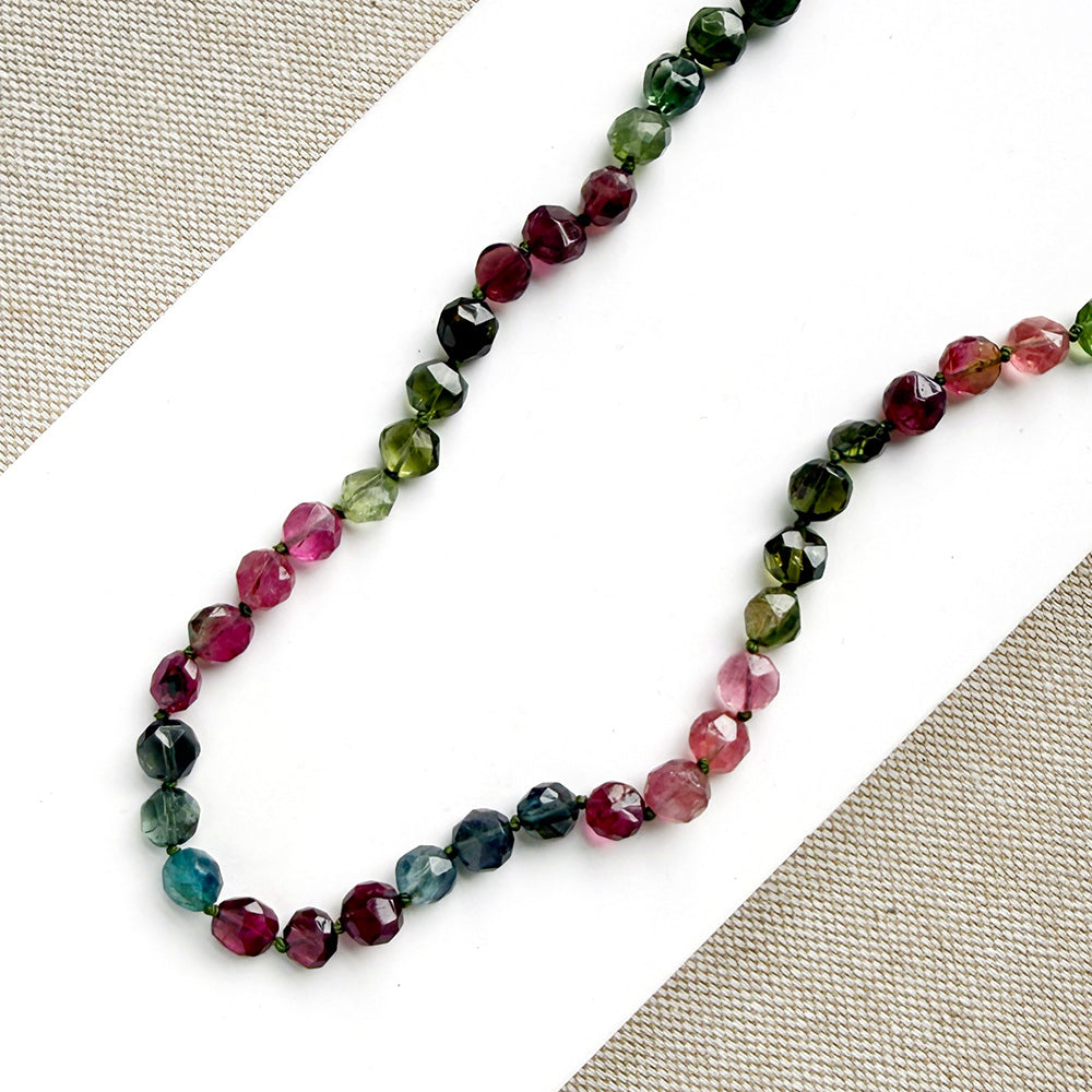 A necklace with faceted Watermelon Tourmaline gemstone beads in a multicolor mix of green, pink, and blue, on a white surface with textured beige fabric in the corners