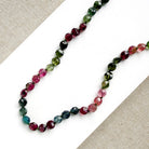 A necklace with faceted Watermelon Tourmaline gemstone beads in a multicolor mix of green, pink, and blue, on a white surface with textured beige fabric in the corners