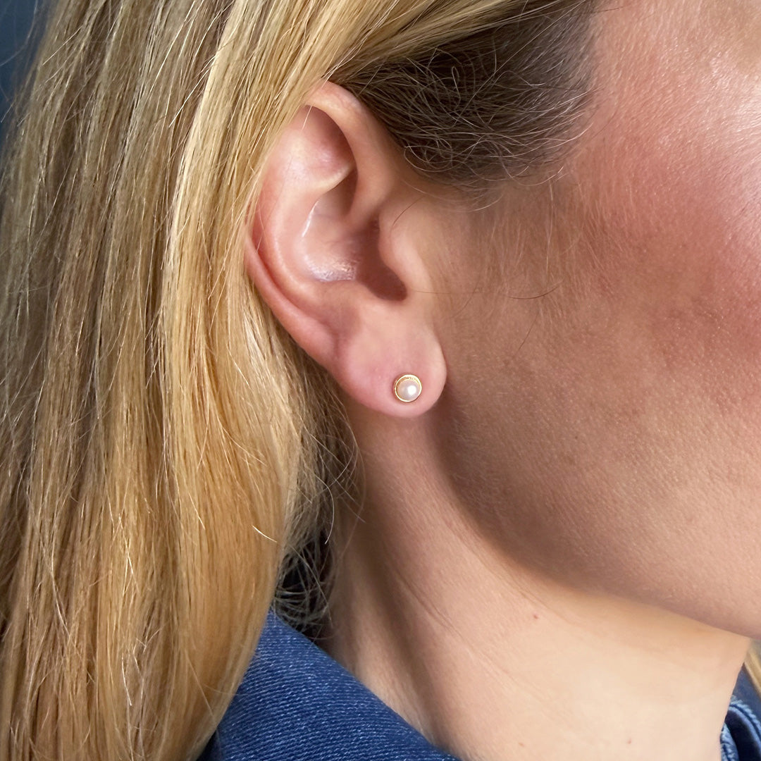 Close-up of a person wearing a pearl and gold stud earring against a blue background