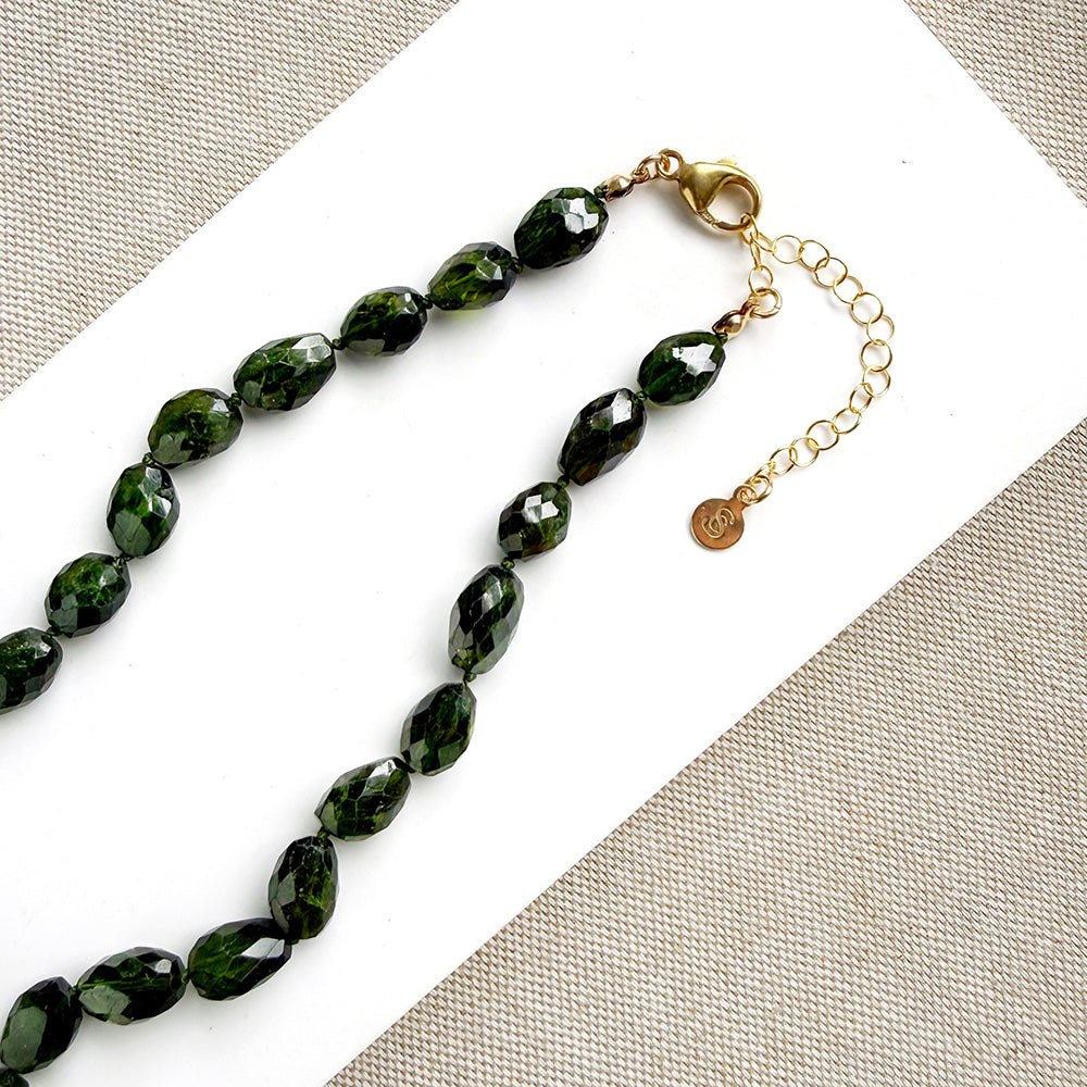 Faceted dark green gemstone beads on a beaded necklace with a gold clasp and extender chain, displayed on a white card over a beige woven fabric background.
