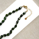 Faceted dark green gemstone beads on a beaded necklace with a gold clasp and extender chain, displayed on a white card over a beige woven fabric background.