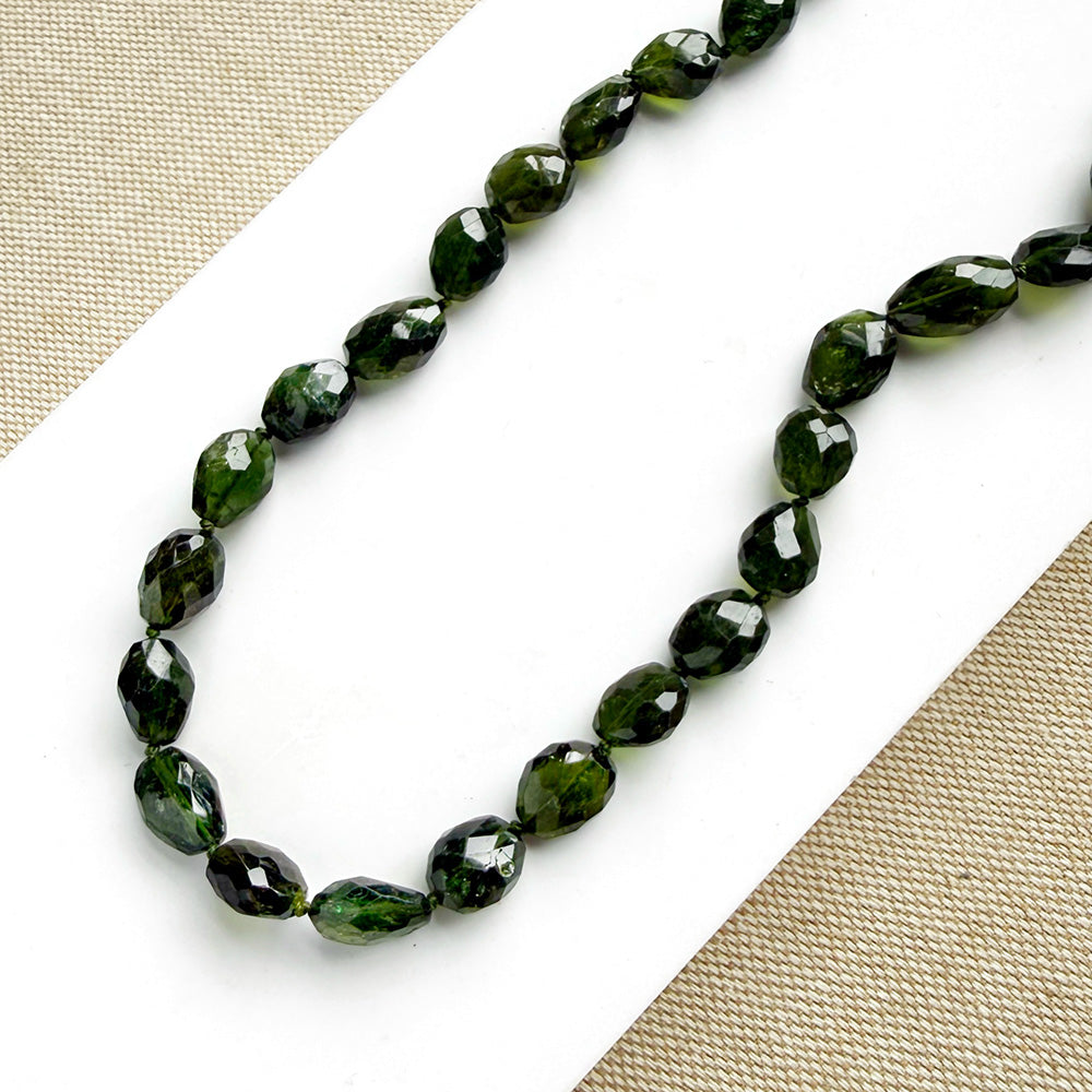 Faceted dark green gemstone beads on a beaded necklace, arranged on a white card over a beige woven fabric background.