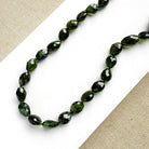 Faceted dark green gemstone beads on a beaded necklace, arranged on a white card over a beige woven fabric background.