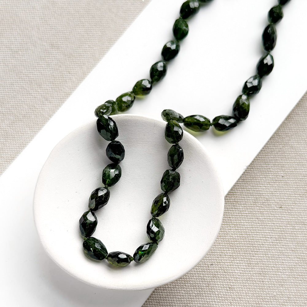 Faceted dark green tourmaline gemstone necklace, draped over a small white ceramic dish on a neutral textured fabric background.