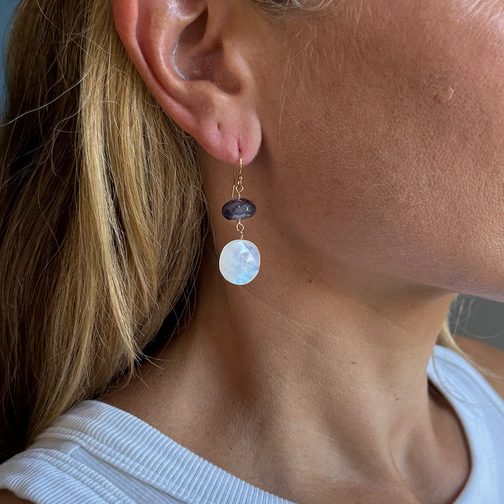 A close-up of a person's ear and neck wearing a dangling earring. The earring has a small dark faceted bead, a gold hook, and a larger round pale blue moonstone drop.