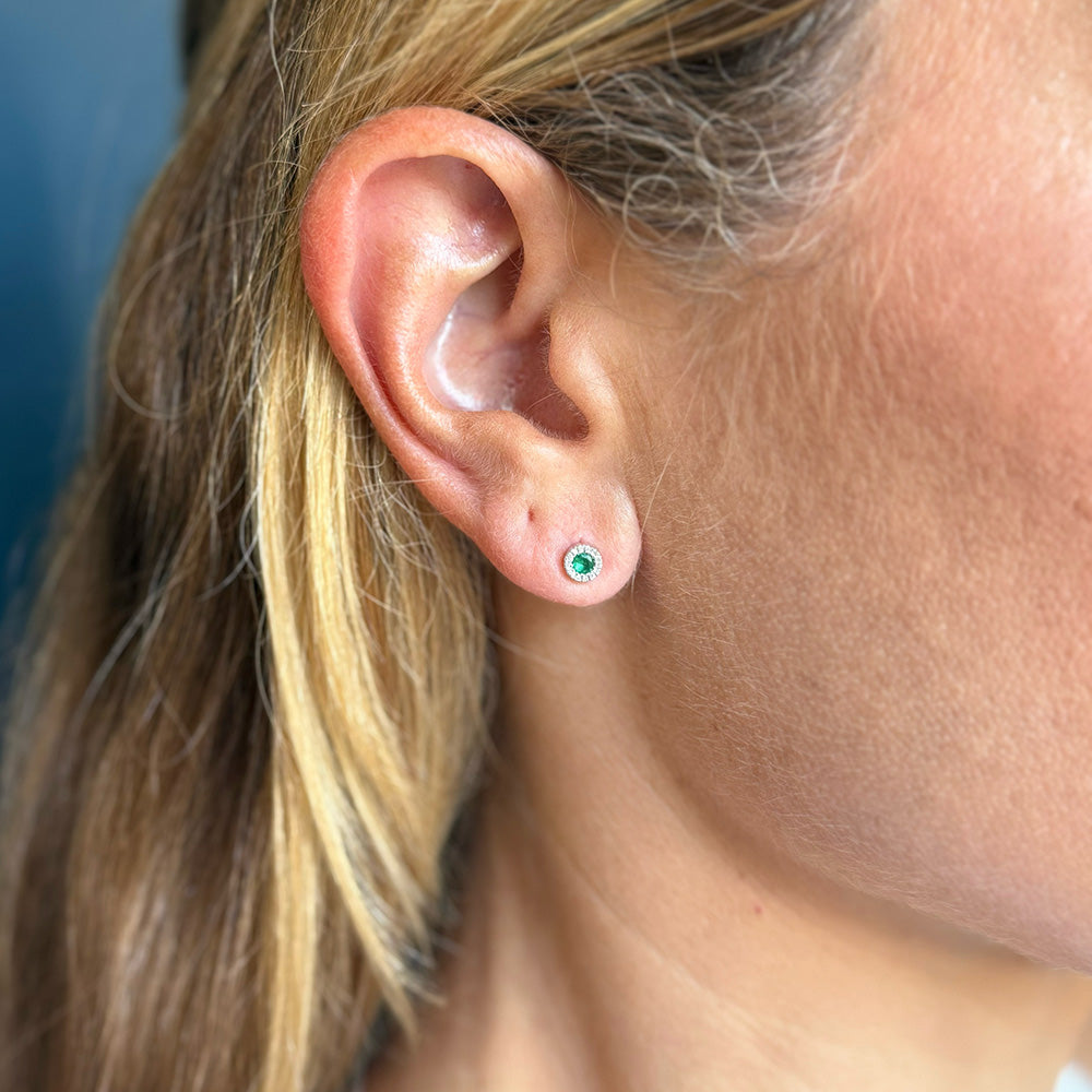 A close-up of a person's ear wearing a small stud earring with a round green emerald in a white gold setting.