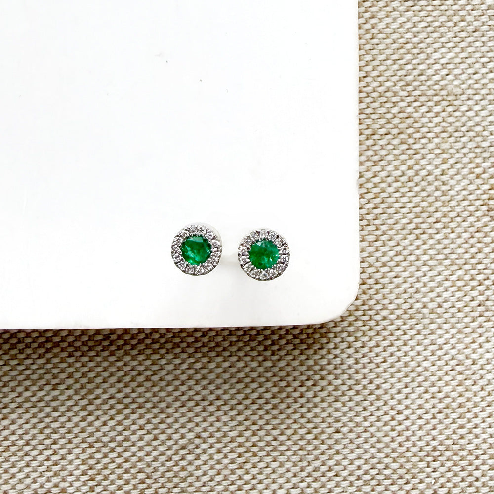 A pair of round emerald and diamond stud earrings, displayed on a white card over a beige woven fabric background.