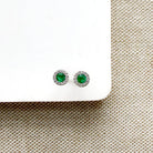 A pair of round emerald and diamond stud earrings, displayed on a white card over a beige woven fabric background.