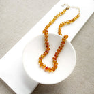 A beaded necklace made of smooth Imperial Topaz with a gold clasp, draped over a small white dish on a textured beige surface.