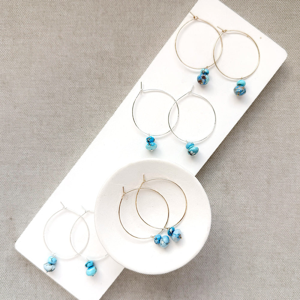 Three pairs of hoop earrings with small Golden Hills Turquoise beads, displayed on a white jewelry stand against a textured gray background.
