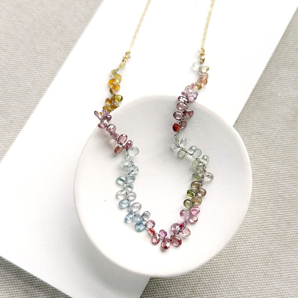 A delicate necklace made of small, tear drop faceted sapphires in a rainbow gradient, displayed on a white circular stand over a textured gray fabric background.