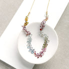 A delicate necklace made of small, tear drop faceted sapphires in a rainbow gradient, displayed on a white circular stand over a textured gray fabric background.
