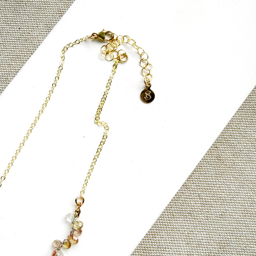 A delicate necklace made of small, tear drop faceted sapphires in a rainbow gradient, displayed on a white card over a textured gray fabric background.