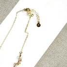 A delicate necklace made of small, tear drop faceted sapphires in a rainbow gradient, displayed on a white card over a textured gray fabric background.