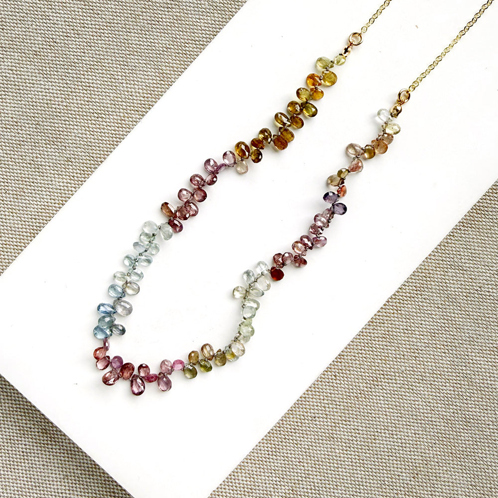 A delicate necklace made of small, tear drop faceted sapphires in a rainbow gradient, displayed on a white card over a textured gray fabric background.