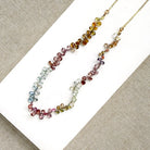 A delicate necklace made of small, tear drop faceted sapphires in a rainbow gradient, displayed on a white card over a textured gray fabric background.
