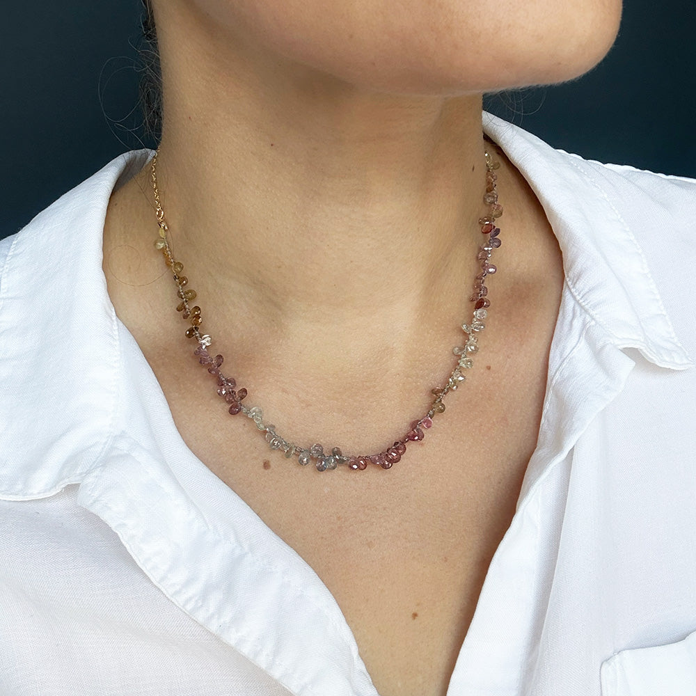 A close-up of a person wearing a delicate necklace made of small, multi-colored sapphires on a gold-toned chain, paired with a white collared shirt.