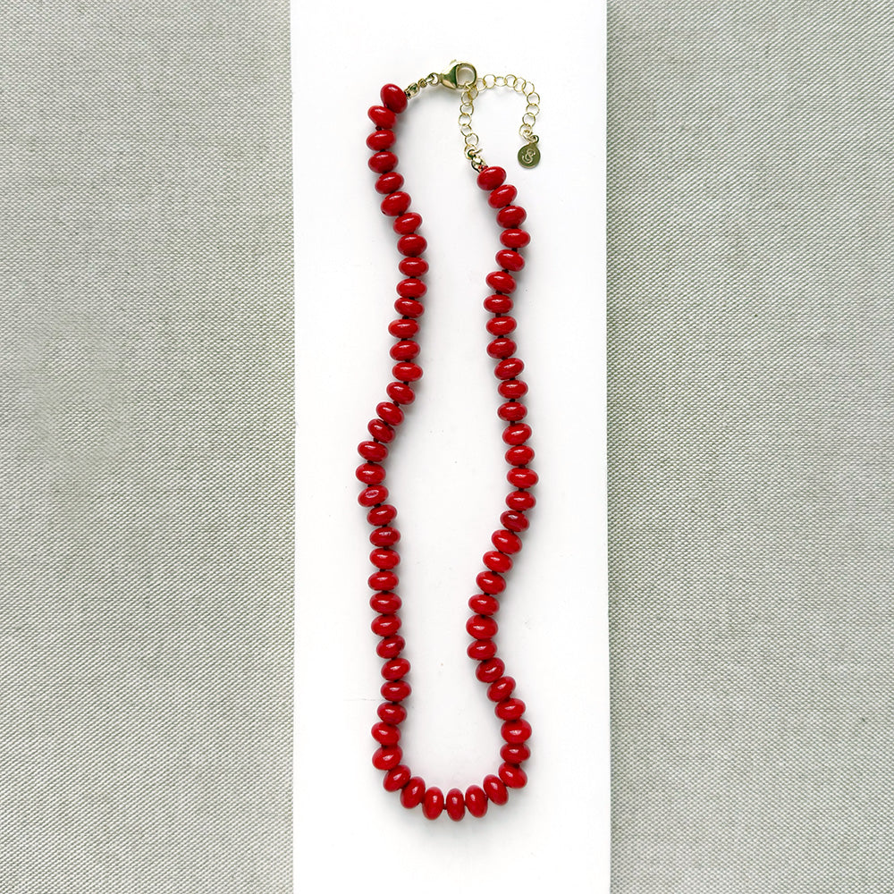 A coral knotted necklace in a rich red color with a gold clasp, displayed on a white ceramic dish over a light gray textured fabric background.