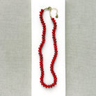 A coral knotted necklace in a rich red color with a gold clasp, displayed on a white ceramic dish over a light gray textured fabric background.