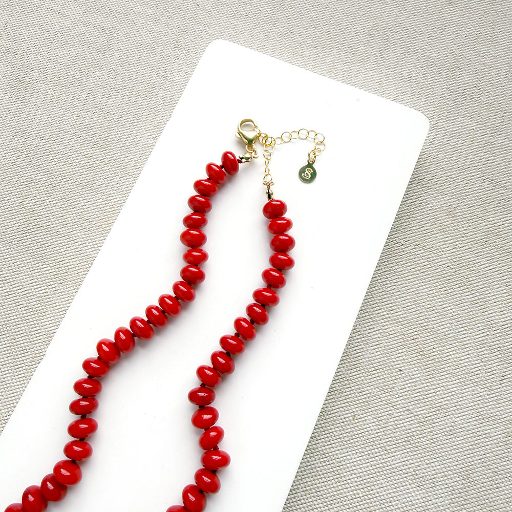 A coral knotted necklace in a rich red color with a gold clasp, displayed on a white ceramic dish over a light gray textured fabric background.
