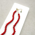 A coral knotted necklace in a rich red color with a gold clasp, displayed on a white ceramic dish over a light gray textured fabric background.