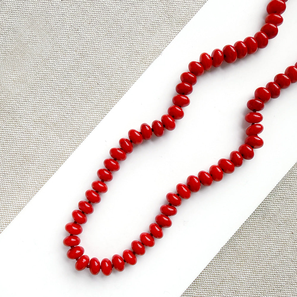 A coral knotted necklace in a rich red color with a gold clasp, displayed on a white ceramic dish over a light gray textured fabric background.