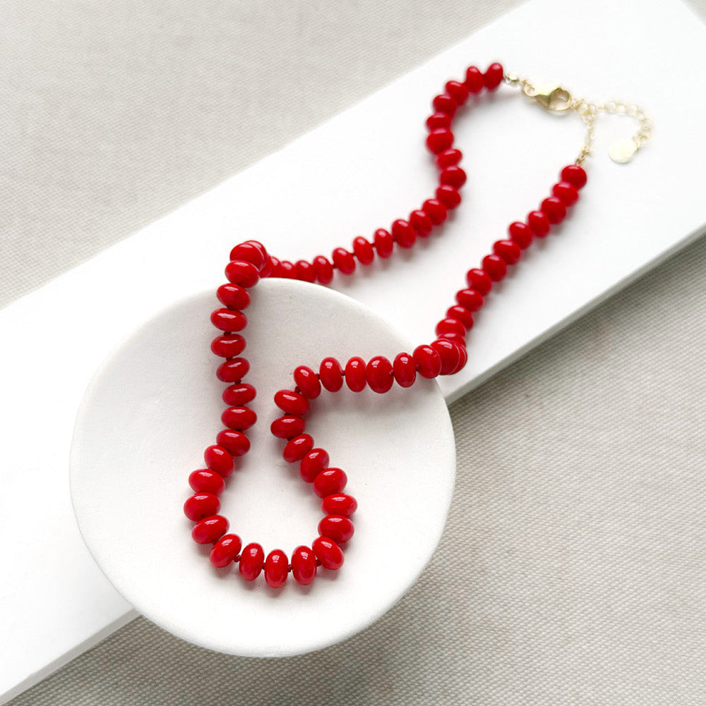 A coral knotted necklace in a rich red color with a gold clasp, displayed on a white ceramic dish over a light gray textured fabric background.