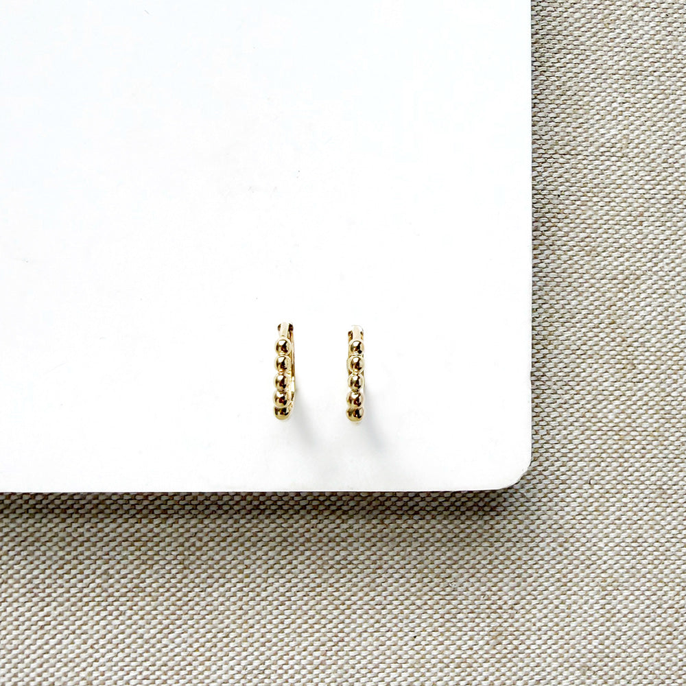 A pair of small gold huggie hoop earrings with a textured, beaded surface, displayed on a white card over a beige woven fabric background.