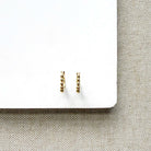 A pair of small gold huggie hoop earrings with a textured, beaded surface, displayed on a white card over a beige woven fabric background.