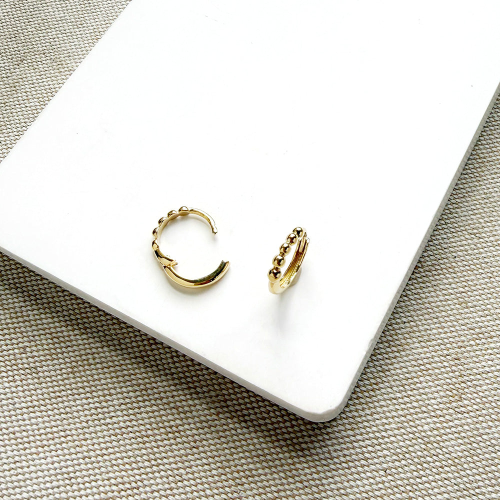A pair of small gold huggie hoop earrings with textured beaded edges, displayed on a white rectangular card over a woven beige fabric background.