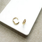 A pair of small gold huggie hoop earrings with textured beaded edges, displayed on a white rectangular card over a woven beige fabric background.