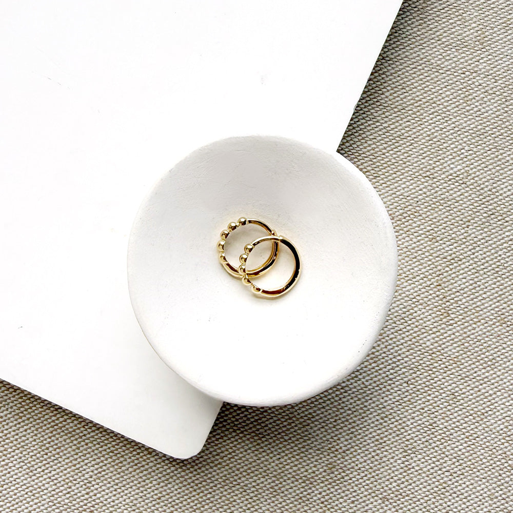 Two delicate gold hoop earrings resting in a small white ceramic dish, set against a textured beige fabric background with a white paper corner.