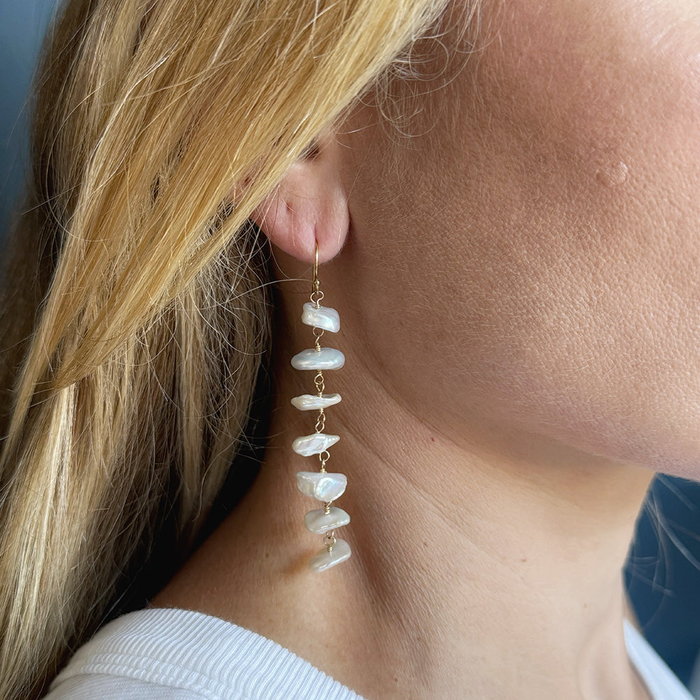 A close-up of a woman's ear wearing a long dangling earring made of irregular white pearls with small gold-tone connectors.