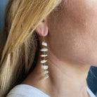 A close-up of a woman's ear wearing a long dangling earring made of irregular white pearls with small gold-tone connectors.
