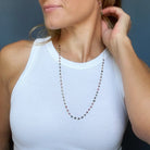 A woman wearing a delicate beaded necklace with watermelon tourmaline gems, paired with a white ribbed tank top against a dark blue-gray background.