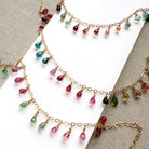 Gold necklaces with dangling teardrop-shaped Watermelon Tourmaline gemstones in pink, green, and teal, displayed on a white card over a beige textured fabric background.