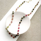 A gemstone necklace with small faceted Watermelon Tourmaline beads in pink, green and blush, draped over a white oval dish on a textured beige fabric background.