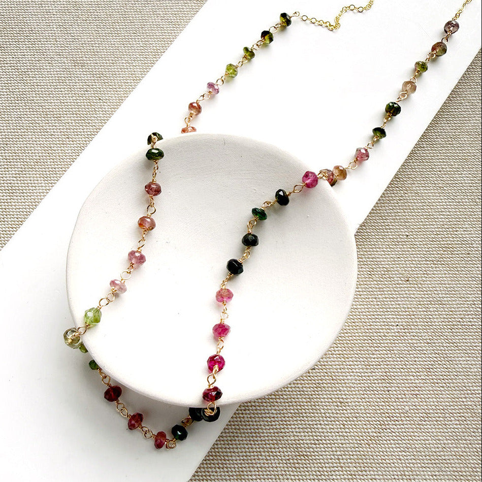 A gemstone necklace with small faceted Watermelon Tourmaline beads in pink, green and blush, draped over a white oval dish on a textured beige fabric background.