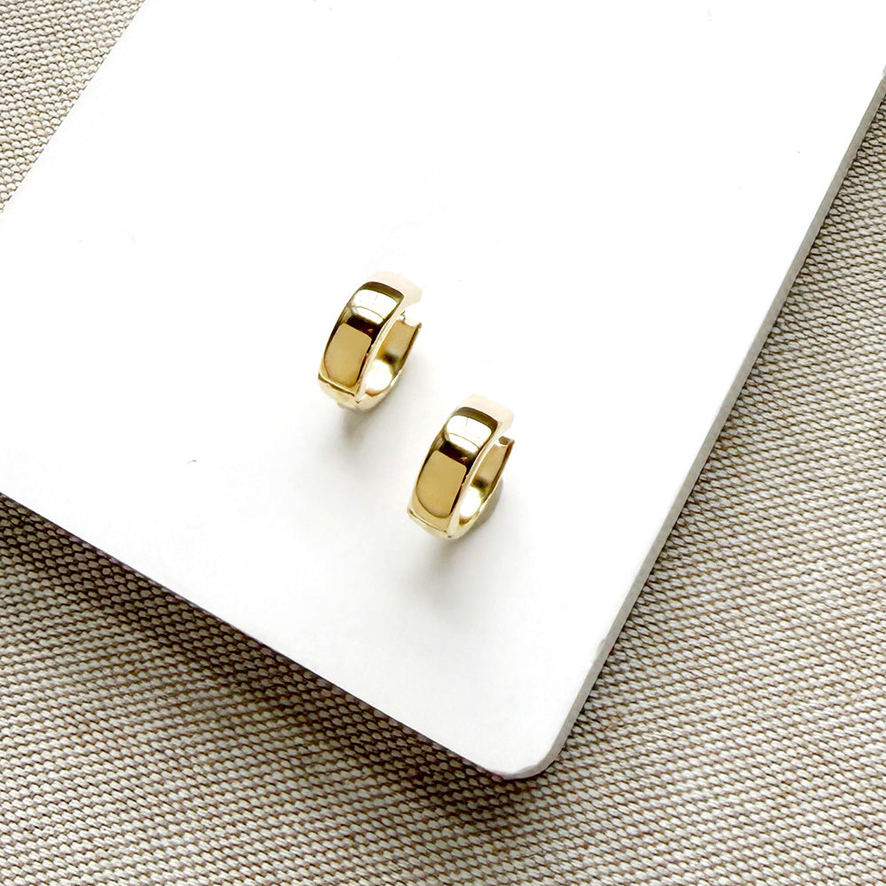 A pair of small gold huggie hoop earrings resting on a white rectangular card over a beige woven fabric background.