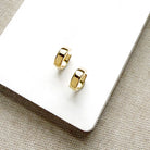 A pair of small gold huggie hoop earrings resting on a white rectangular card over a beige woven fabric background.